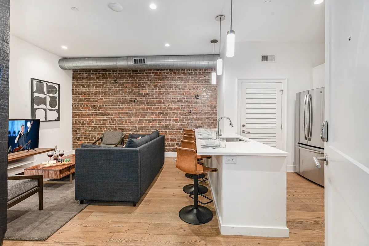 A spacious urban loft interior featuring an exposed brick wall, a modern couch arranged in a welcoming living space, a kitchen island perfect for gatherings, and a television mounted for entertainment. The setting exudes a stylish, comfortable city ambiance, ideal for relaxing or entertaining friends after a night out.