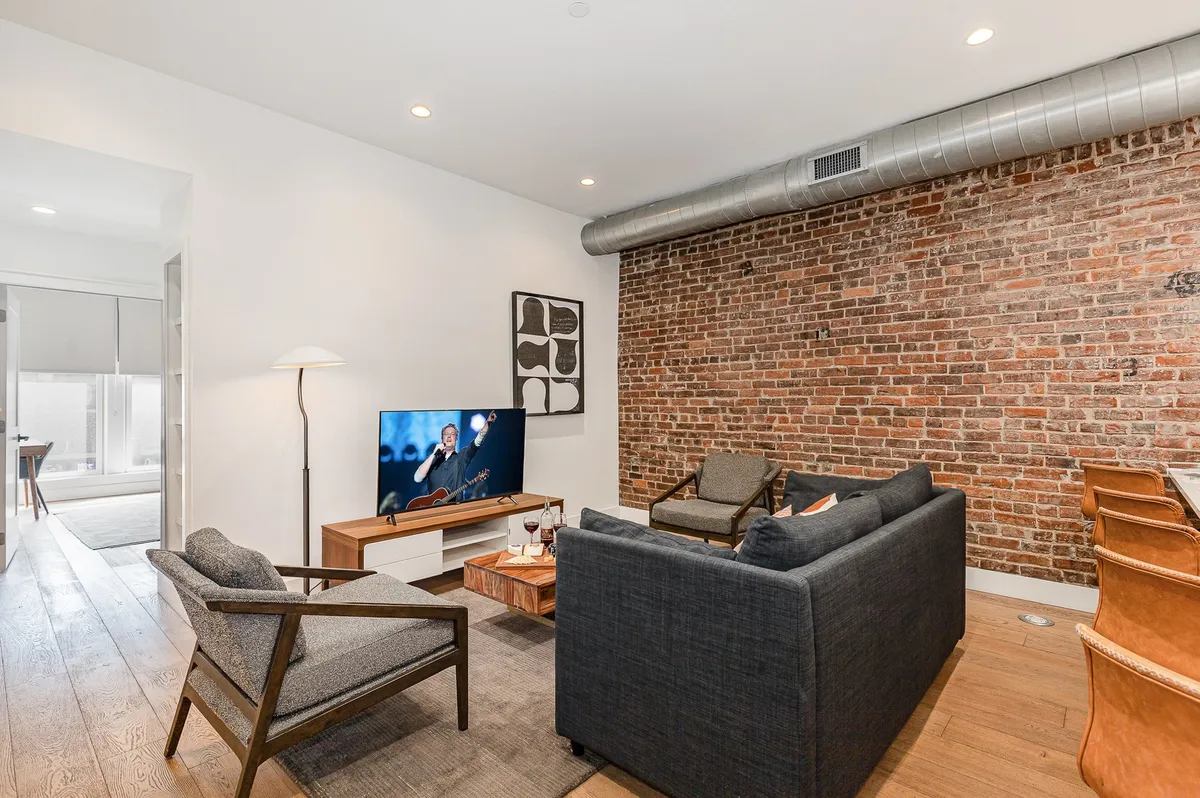 A cozy living room scene with an exposed brick wall, a stylish sofa, comfortable armchairs, a large television, and a modern lighting fixture. The space radiates warmth and is ideal for entertainment and intimate conversations, evoking the spirit of camaraderie and togetherness.