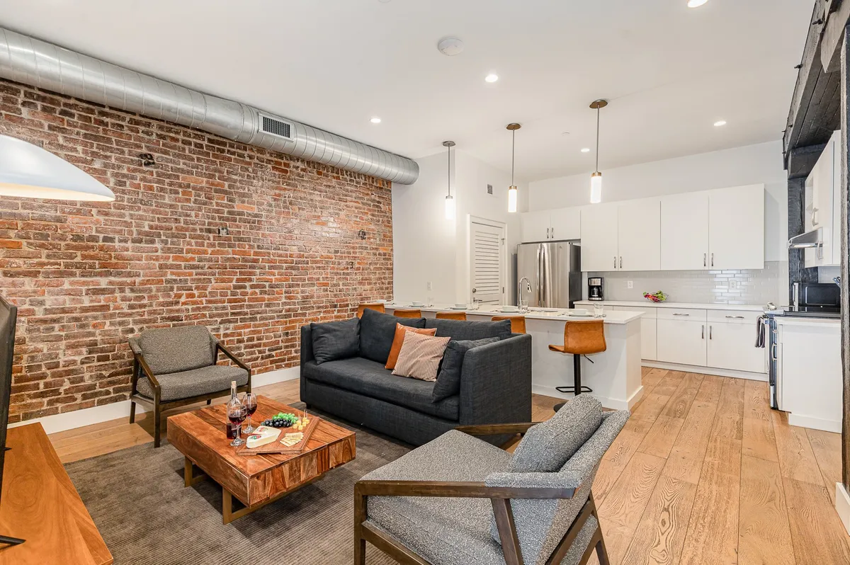 A welcoming open-concept living space featuring a warm exposed brick wall, sleek modern kitchen with a large island, and a plush seating area arranged around a rustic wood coffee table. The scene exudes coziness and urban charm, perfectly suited for social gatherings, relaxing evenings, or festive group dinners.