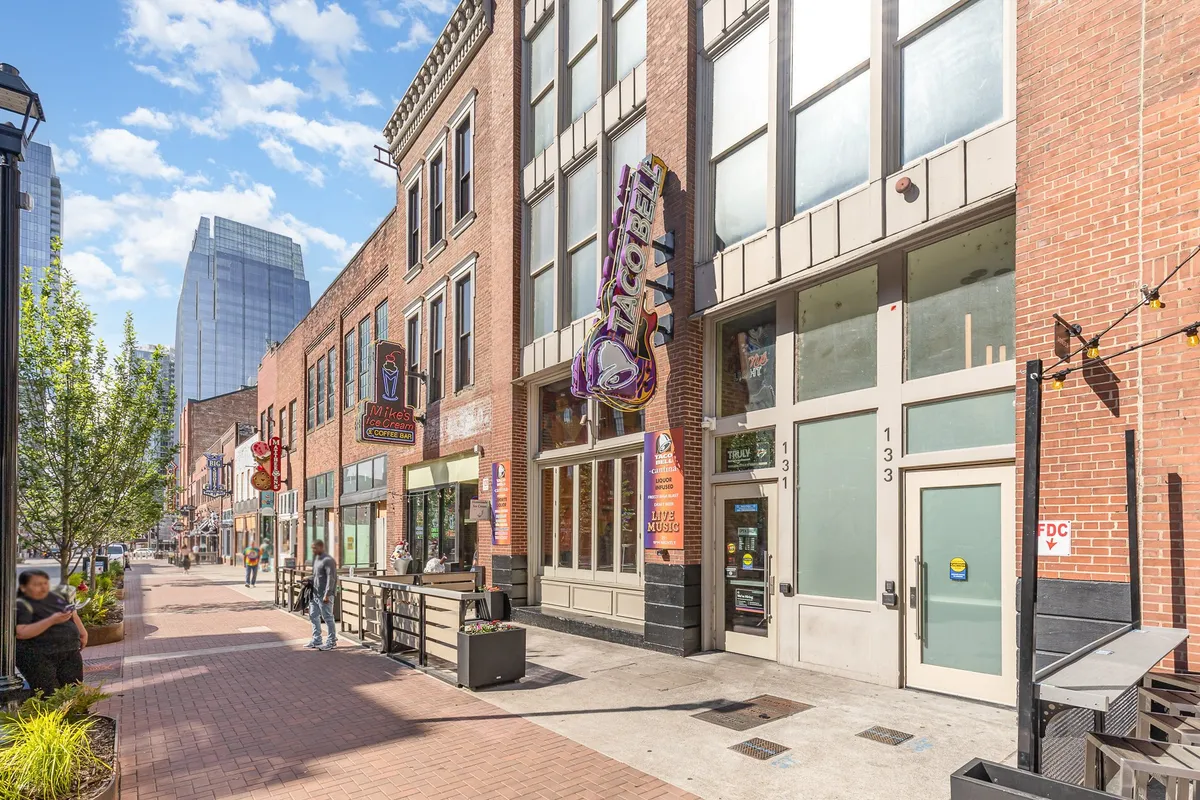 An outdoor urban scene bustling with people, restaurant facades, and outdoor seating under sunny skies. The Taco Bell sign and lively pedestrian activity suggest a vibrant, walkable neighborhood filled with dining and social opportunities.