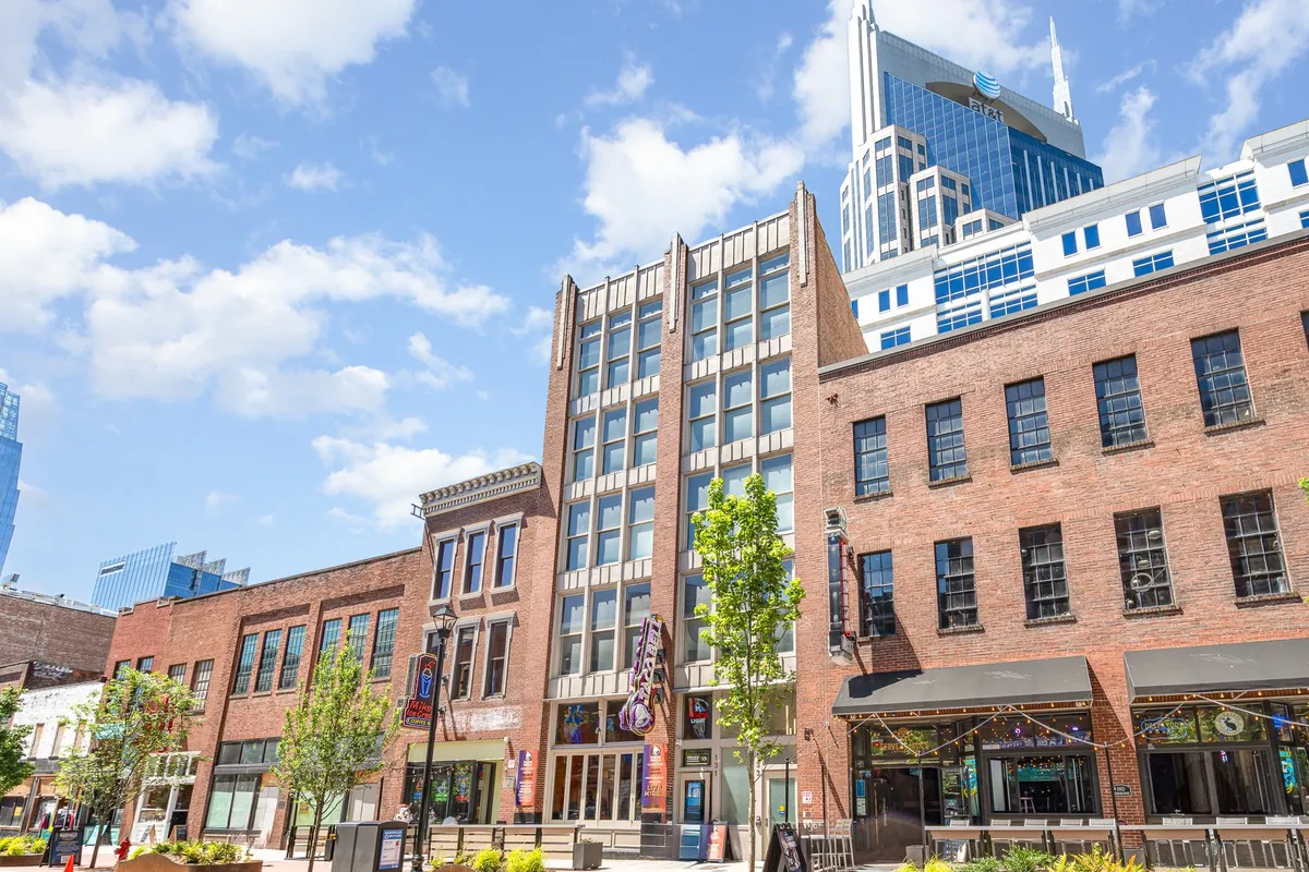 A vibrant city scene featuring a historic brick building juxtaposed with a modern skyscraper, green trees, and an inviting outdoor seating area bustling with people dining, shopping, and socializing.