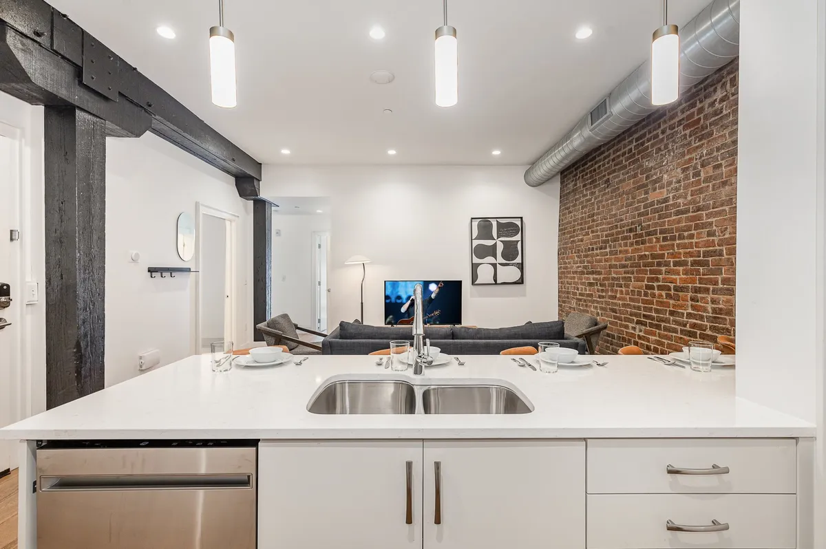 A welcoming and contemporary loft living space featuring a kitchen island with bar seating, exposed brick wall, TV, and a plush sofa. The warm lighting and inviting layout make it easy to imagine celebrating or unwinding after a busy day downtown.