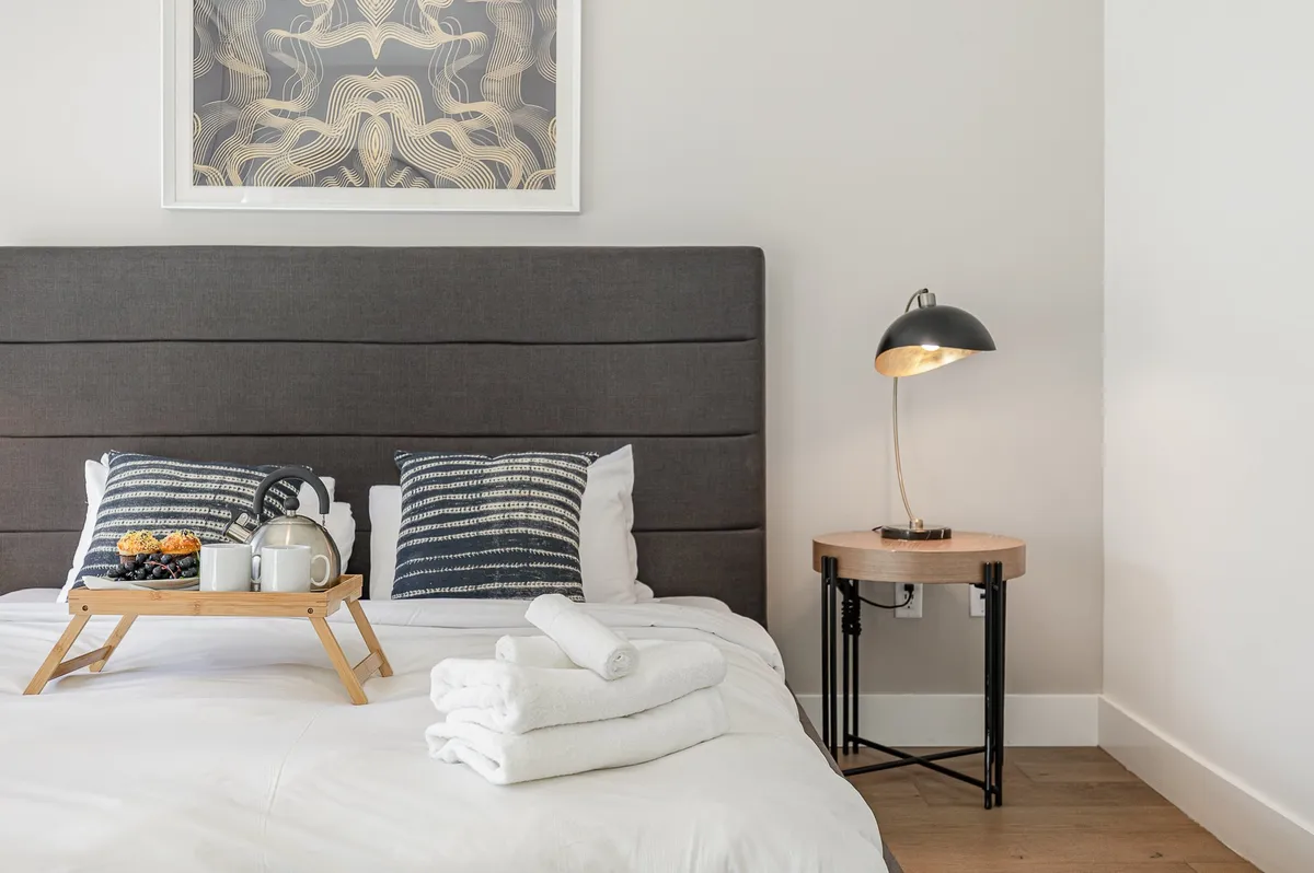 A serene bedroom features a plush bed adorned with decorative pillows, a breakfast tray set with tea and fruit, tasteful artwork hanging above, and a modern lamp on the nightstand. The soft lighting and cozy textures create an inviting space for rest and rejuvenation.