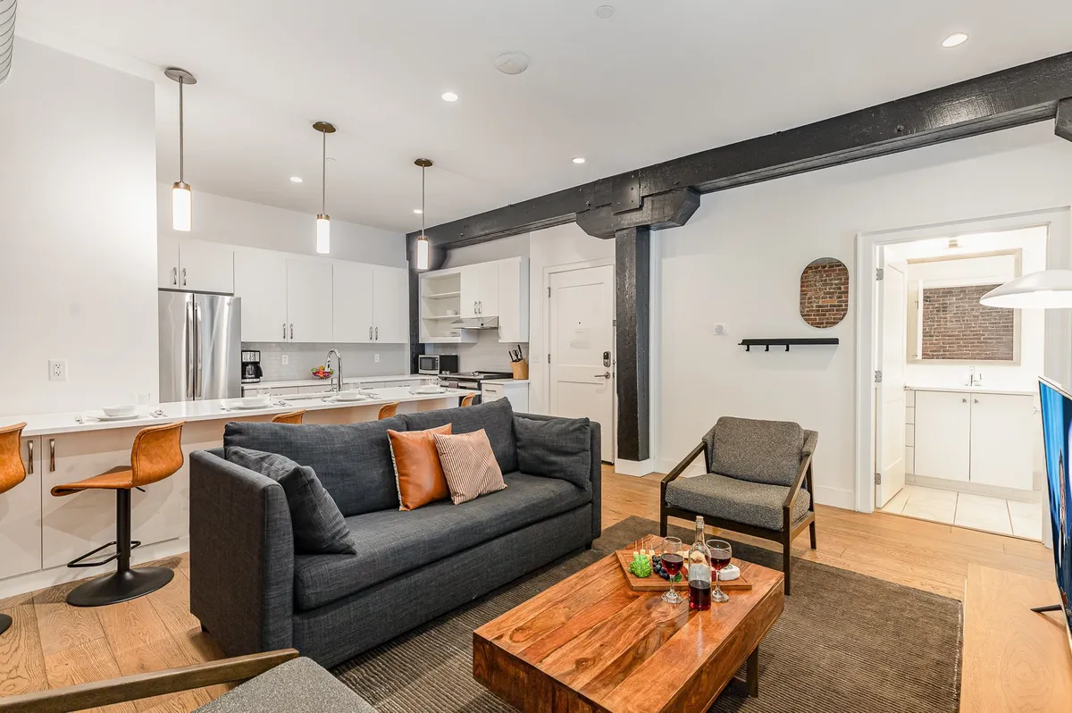 This image features a cozy, contemporary open-concept living space with a comfortable sofa, a modern coffee table, a kitchen island with bar stools, and artwork on the wall. The layout creates an inviting area for socializing, relaxing, and entertaining, showcasing spaces where guests can lounge, enjoy snacks, or play games.